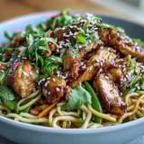 A steaming bowl of Sesame Chicken Noodle Bowl garnished with sesame seeds, fresh cilantro, and bright lime wedges.