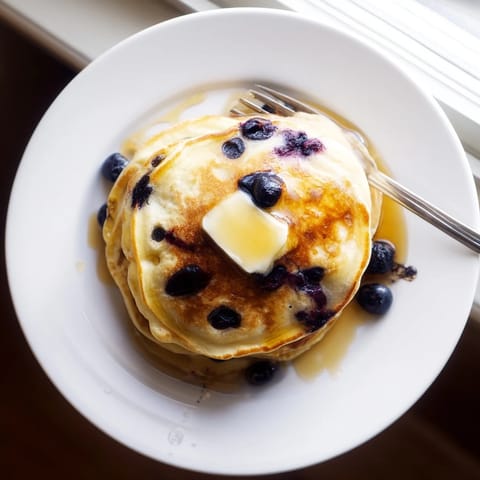 Freshly cooked blueberry pancakes stacked high, topped with melting butter and warm maple syrup for a cozy breakfast.
