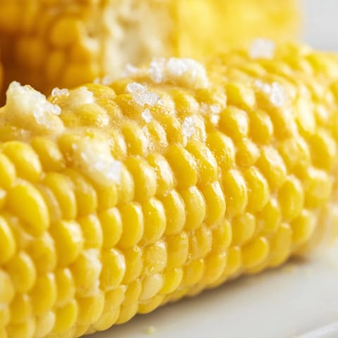 Tender boiled corn on the cob, slathered with creamy butter and a pinch of salt on a plate.