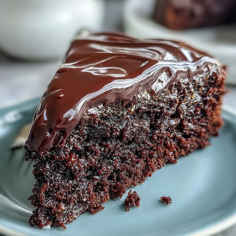 A slice of Easy Chocolate Fudge Cake on a white plate reveals a dense, squidgy crumb, served alongside a cup of hot coffee.